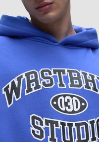 Niebieska bluza z kapturem z haftowanym napisem "WRSTBH" oraz "STUDIO" w czerni i bieli. Posiada dużą kieszeń typu kangur na przodzie oraz kaptur ze sznurkiem.
