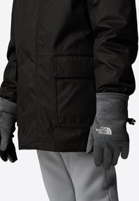 Black insulated jacket with a pocket and gray gloves. Gloves have a textured finish and logo. Model wears light gray pants.
