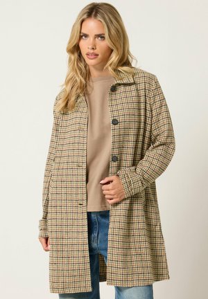 Femme blonde portant un manteau à motif pied-de-poule beige et marron par-dessus un t-shirt beige et un jean bleu sur un fond clair uni.
