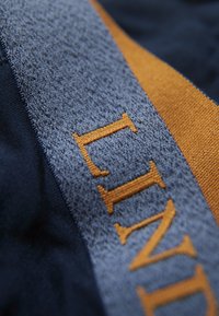 Tessuto blu marina con una fascia grigia testurizzata; presenta una scritta arancione bold con una leggera lucentezza. Le cuciture sono visibili sui bordi.