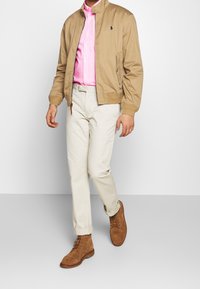 Tan bomberjacka över en rosarandig skjorta, krämfärgade byxor och bruna mockaskor med snörning. Avslappnad, modern outfit med rena linjer.