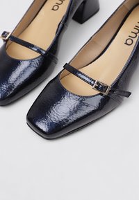 Talons en cuir verni marine avec un bout carré, une sangle sur le coup de pied et un petit talon carré. Texture lisse avec une finition brillante.