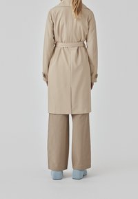 Beige trenchcoat met een riem, een split aan de achterkant en lange mouwen. Gecombineerd met brede beige broek en blauwe hakken.