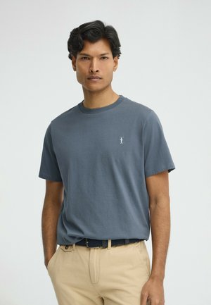 Homme portant un t-shirt bleu uni avec un petit logo blanc sur la poitrine, un pantalon beige et une ceinture noire tressée, debout les mains dans les poches.