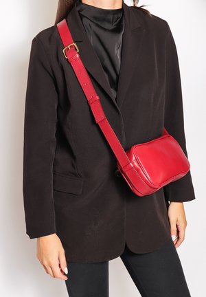 Borsa a tracolla rossa in pelle con tracolla regolabile, forma rettangolare, superficie liscia e una leggera lucentezza. Indossata sopra un blazer nero.
