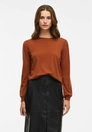 Haut à manches longues couleur rouille avec un col rond et des fronces subtiles au niveau des manches, associé à une jupe en cuir noir. Texture lisse.