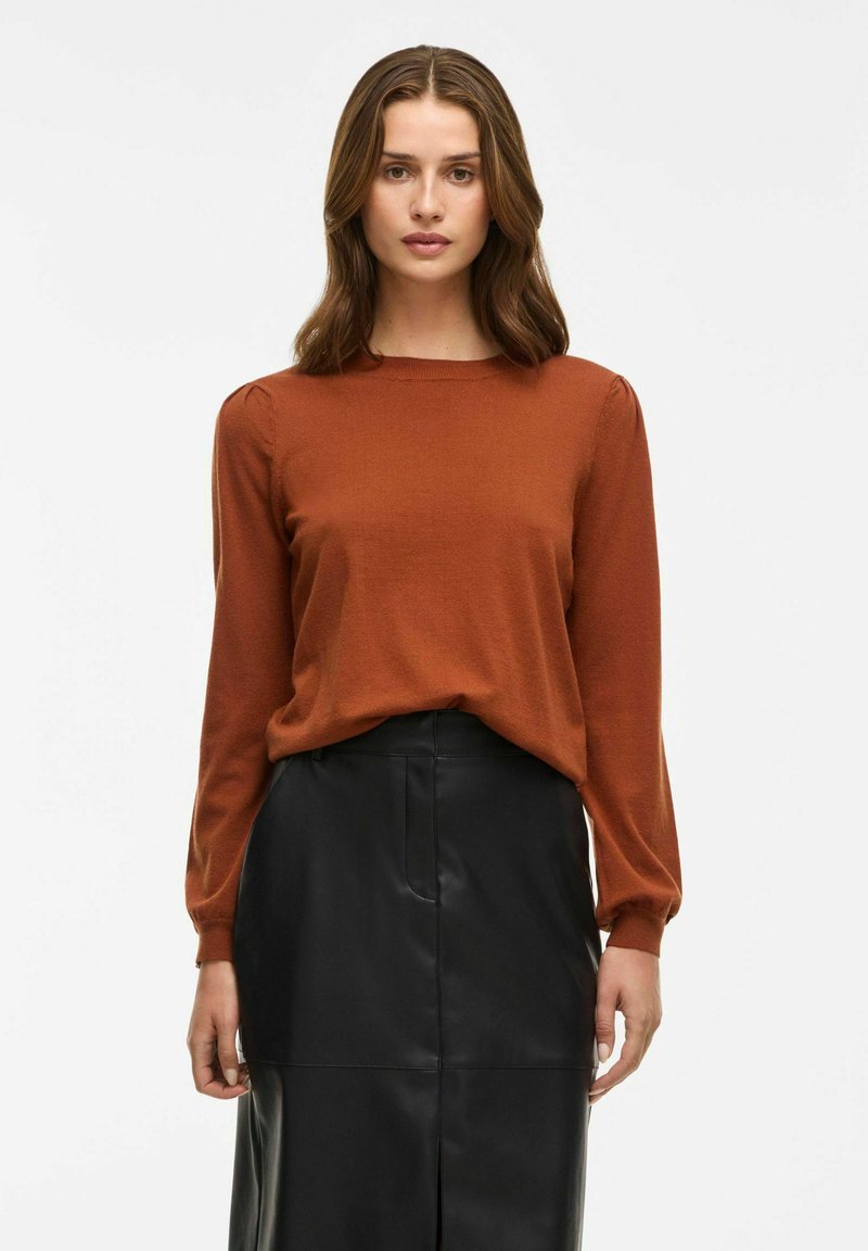 Haut à manches longues couleur rouille avec un col rond et des fronces subtiles au niveau des manches, associé à une jupe en cuir noir. Texture lisse.
