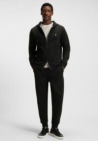 Uomo che indossa una felpa nera con zip, pantaloni jogger neri, t-shirt bianca e sneakers nere, in piedi con le mani in tasca su uno sfondo neutro.