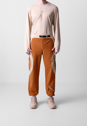 Osoba ubrana w beżową koszulę z długim rękawem i pomarańczowe spodnie cargo z beżowymi panelami po bokach, pasek i beżowe trampki na jednolitym jasnym tle.