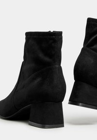 Botas de tobillo de ante negro con tacón cuadrado, que presentan un caña alta y una textura suave, mostrando un diseño minimalista y líneas limpias.