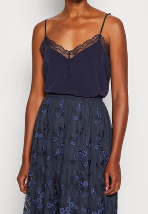 Kvinde iført en navy camisole med blondekant og knapper, kombineret med et mørkt gennemsigtigt skørt med blå blomsterbroderi.