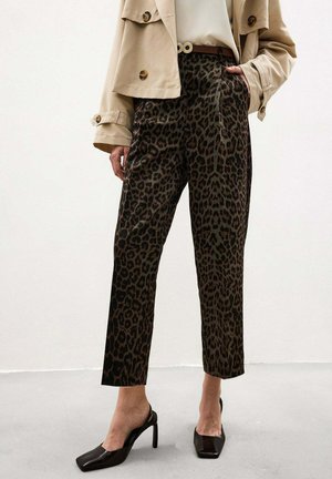 Žena nosí béžovú krátku bundu, vysoké nohavice s leopardím vzorom, čierne slingback lodičky, stojí s jednou rukou v vrecku.