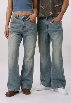 Δύο άτομα με φαρδιά, ξεθωριασμένα μπλε τζιν και τα χέρια στις τσέπες· το ένα φοράει καφέ μπότες, το άλλο λευκά αθλητικά παπούτσια.