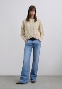 Pull crème en maille torsadée à col en V, coupe oversize, porté sur un haut léger. Jean bleu à jambes larges et ballerines pointues marron complètent la tenue.