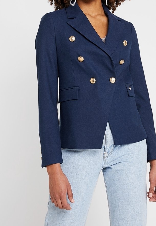 Navy kaksoisnapitettu bleiseri, jossa on kultaiset napit, muotoillut kaulukset ja etutaskut, yhdistettynä vaaleansinisisiin farkkuihin.