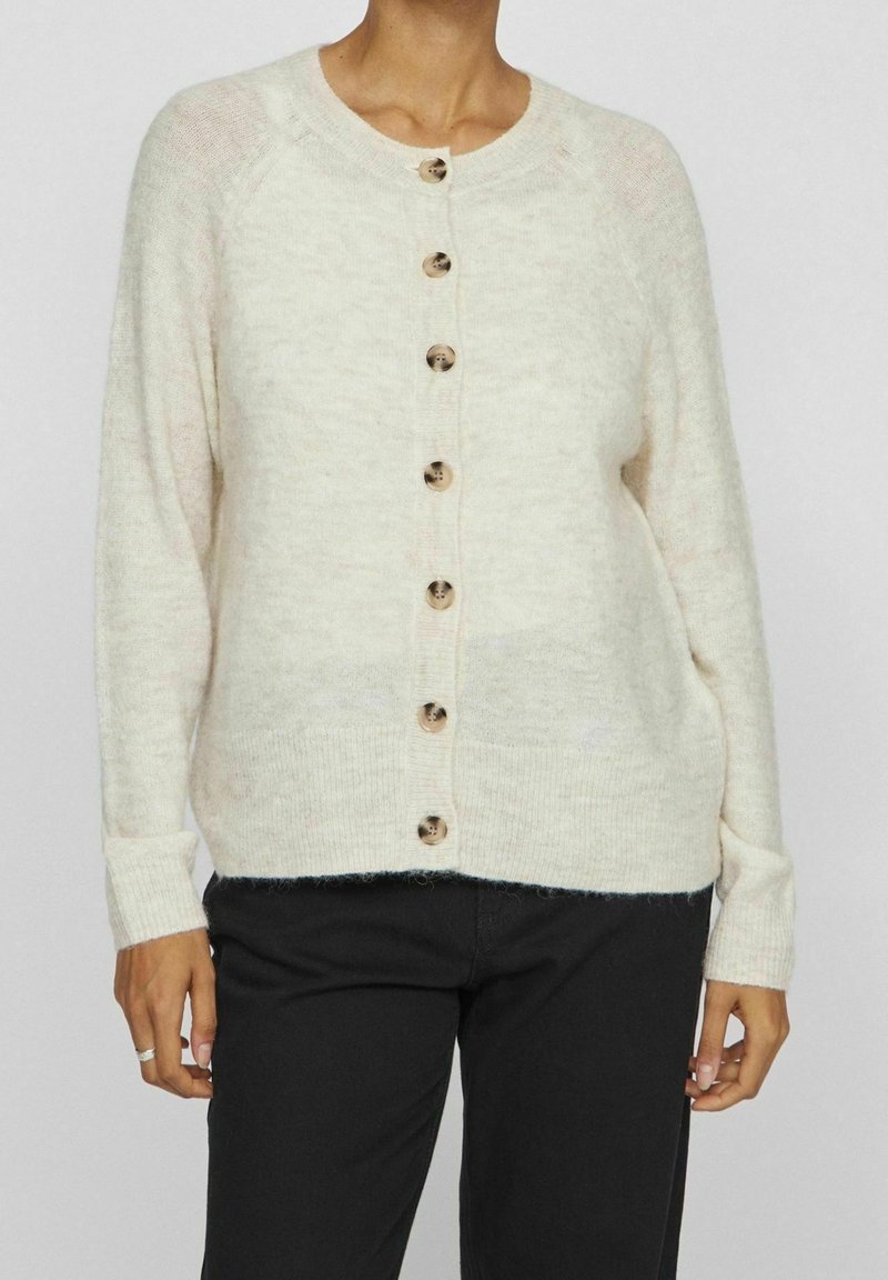 Femme portant un cardigan boutonné de couleur crème à manches longues et un pantalon noir, debout devant un fond uni.