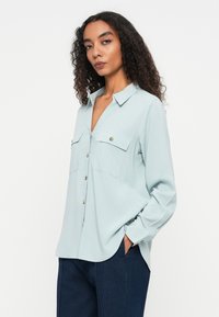 Jasnoniebieska, długorękawowa koszula z kołnierzykiem, z przodu zapinana na guziki, z dwoma kieszeniami na piersi. Miękki, gładki materiał o luźnym kroju.