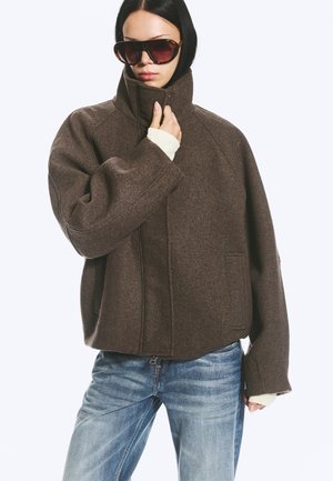 Femme portant une veste en laine marron oversize, un jean bleu clair et de grandes lunettes de soleil foncées, tenant le col de la veste d'une main.