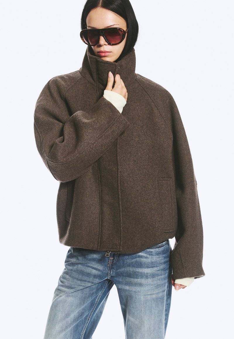 Femme portant une veste en laine marron oversize, un jean bleu clair et de grandes lunettes de soleil foncées, tenant le col de la veste d'une main.