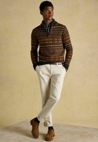 Kötött pulóver színes geometrikus mintával, sötétzöld gallérral, krémszínű nadrággal és barna velúr cipővel párosítva, laza fazonban.