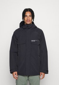 Junger Mann mit geflochtenem Haar, der eine schwarze Winterjacke mit Kapuze und Taschenklappen sowie grüne Hosen trägt, steht vor einem schlichten Hintergrund.