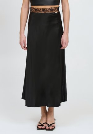 Falda de satén negra con una cinturilla de encaje, forma en A, textura suave y aberturas laterales, combinada con sandalias negras de tiras.