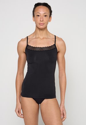 Kobieta z zaplecionymi włosami, ubrana w czarny top na ramiączkach z koronkową lamówką i czarną bieliznę, stojąca na jednolitym jasnym tle.