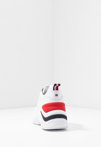 Baskets de sport blanches avec une tige en maille, accent rouge sur le talon, détail à rayures bleues et rouges sur la languette, et marque "TOMMY HILFIGER".