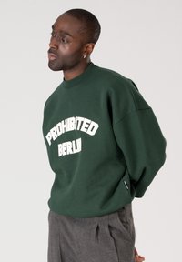 Homme portant un sweat-shirt vert foncé avec l'inscription « PROHIBITED BERLIN », pantalon gris, regardant vers la gauche sur un fond uni.