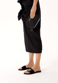 Sorte ruskindsklipklappere med gulddetalje i logo, kombineret med en sort quiltet clutchtaske, holdt i hånden mod en ensfarvet baggrund.