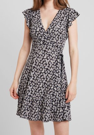 Frau trägt ein schwarzes Wickelkleid mit weißem und gelbem Gänseblümchenmuster und gerafften kurzen Ärmeln, steht mit entspannten Armen.