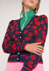 Kvinna bär en blå kofta med röd blommönster, pärlknappar, grön zip-up topp under, rosa byxor, guldringar och stora hoopsörhängen.