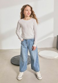 Maglietta grigia chiara a maniche lunghe con un design testurizzato, abbinata a jeans in denim blu a gamba larga e sneakers alte bianche. Outfit semplice e casual.