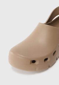 Beige klump med en rund tå, lavet af glat, holdbart materiale. Har sideventilationshuller og en rygstrop for en sikker pasform.