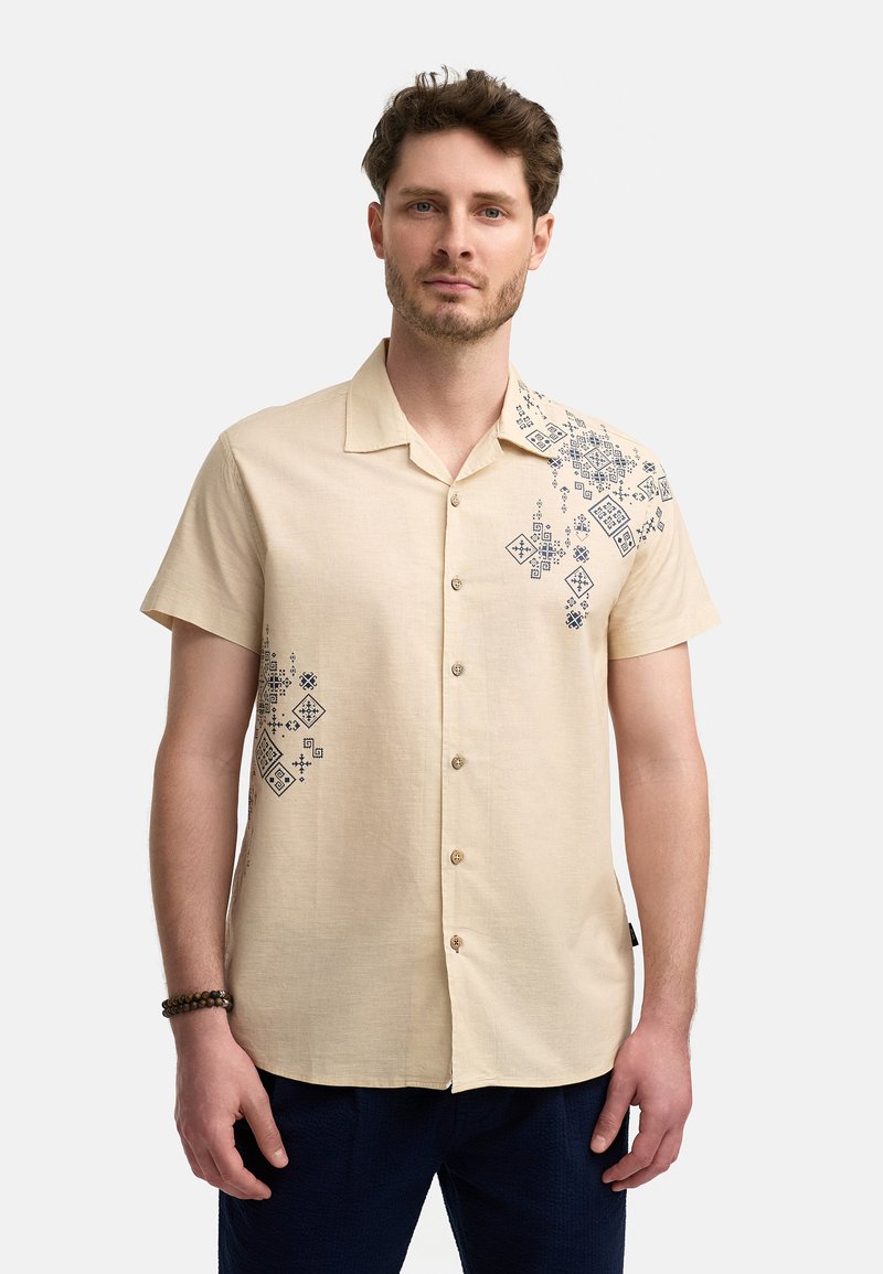 Lyhythihainen beige paita, joka on valmistettu kevyestä kankaasta. Paidan vasemmassa olkapäässä on sinisiä geometrisiä kuvioita ja rantakuvioinen etu.