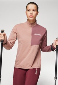 Terrex zip-up topp i lys rosa og burgunder med brystlomme; tettsittende design med lange ermer. Kombinert med matchende burgunder leggings.