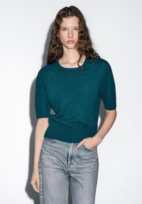 Türkis gestrickter Pullover mit kurzen Ärmeln, rundem Halsausschnitt und geripptem Saum, kombiniert mit hellblauen, gerade geschnittenen Jeans. Glatte Textur, minimales Design.