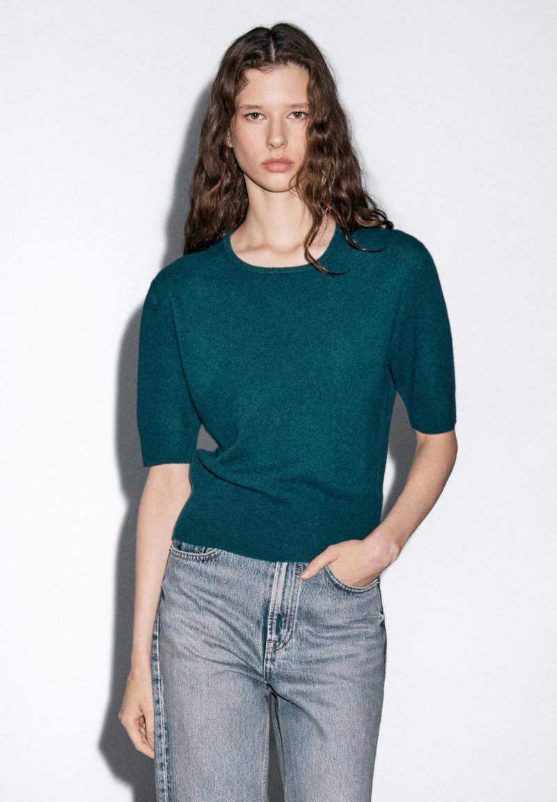 Türkis gestrickter Pullover mit kurzen Ärmeln, rundem Halsausschnitt und geripptem Saum, kombiniert mit hellblauen, gerade geschnittenen Jeans. Glatte Textur, minimales Design.