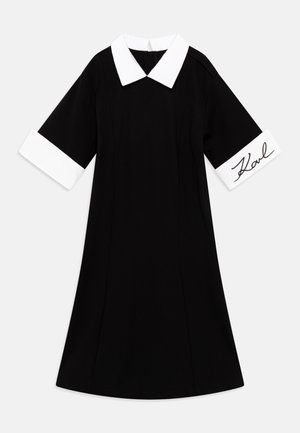 Schwarzes Kleid mit weißem Kragen und Manschetten. Die Manschetten sind mit einer Signatur in schwarzer Schrift verziert. A-Linien-Schnitt, kurze Ärmel, klares Design.