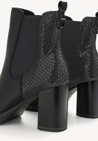Černé kotníkové boty na vysokém podpatku se panely z hadí ků�že, elastickými bočními vsadkami a poutky na bílém pozadí.