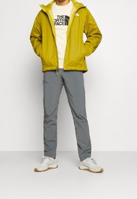Chaqueta impermeable amarilla con capucha, pantalones grises y zapatillas blancas. La chaqueta cuenta con un cierre de cremallera y un logo en el pecho.