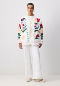 Valge, tikanditega jakk, millel on värvikad lille- ja abstraktsed mustrid, paelaga kinni tõmmatav ja kahe ees oleva taskuga. Kombineeritud valgete flared-pükstega.