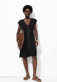 Robe noire à bouton sur le devant avec des manches courtes, tissu texturé et taille cintrée. Accessoirisée avec un sac à franges marron et des sandales marron.