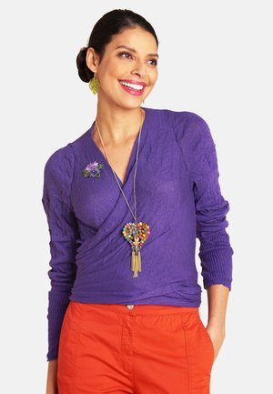 Lächelnde Frau, die einen violetten Wickelstrickpullover mit einer floralen Brosche, eine bunte Herz-Anhänger-Halskette und orangefarbene Hosen trägt, mit der Hand in der Tasche.