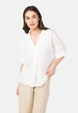 Blusa bianca a maniche corte con scollo a V, con risvolti, in tessuto leggero e vestibilità comoda, abbinata a pantaloni beige.