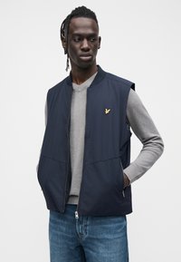 Sötétkék ujjatlan dzseki, elöl cipzáros, arany sas logóval a mellkason, világosszürke pulóver és kék farmer felett viselve.