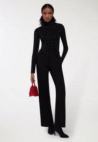Maglione nero a collo alto con decorazioni argento, pantaloni neri a gamba larga e una piccola borsa rossa. La modella indossa stivaletti neri alla caviglia abbinati.