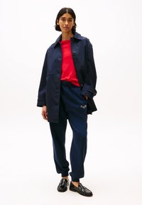 Cappotto impermeabile blu navy con grandi bottoni, camicia rossa a maniche lunghe, pantaloni della tuta blu navy e mocassini in pelle nera con dettagli testurizzati.