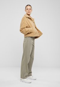 Giacca in finta shearling beige con colletto e chiusura a bottoni, abbinata a pantaloni a gamba larga verdi oliva e sneakers bianche.