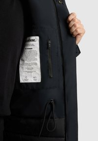 Interiorul unei jachete negre, prezentând o pungă cu fermoar, eticheta de întreținere cu text și un tiv cu șnur. Textură netedă; fără modele sau embellishe. 